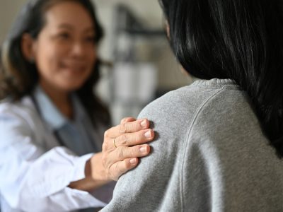 An aged Asian female doctor touching shoulder to comfort and support her patient. A young Asian female patient is being reassured by her doctor. close-up image