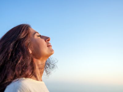 Young woman breathing fresh air with closed eyes and serene expression, enjoying freedom and tranquility under a clear blue sky